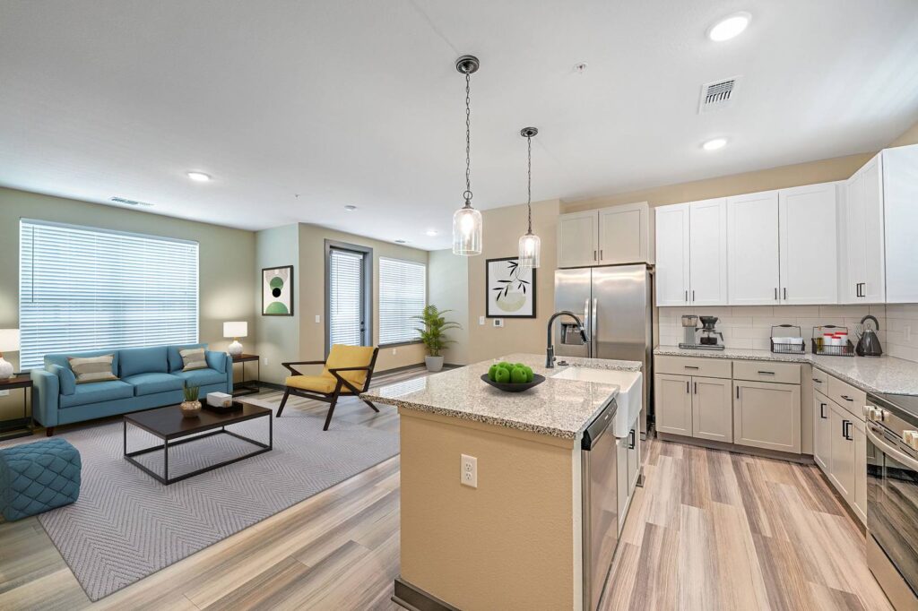 Modern open-plan living room and kitchen with light wood flooring, a blue sofa, and gray kitchen cabinets. A kitchen island with hanging lights sits in the foreground.