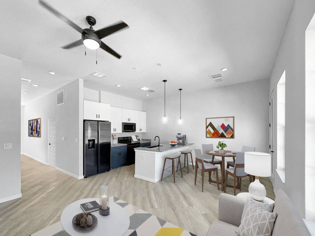 Modern open-concept living space with a kitchen, dining area, and seating. Neutral walls, light wood flooring, ceiling fan, and wall art. White cabinets and black appliances in kitchen.