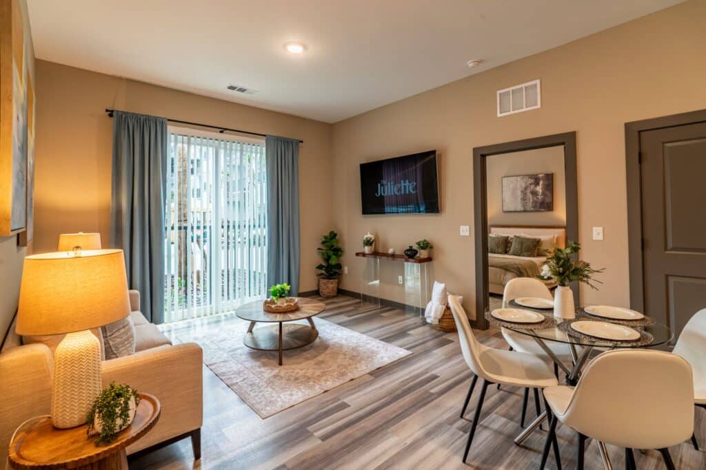 Modern living and dining area with a gray sofa, coffee table, TV, dining set for four, indoor plants, and large window with curtains. Light wooden flooring and beige walls.