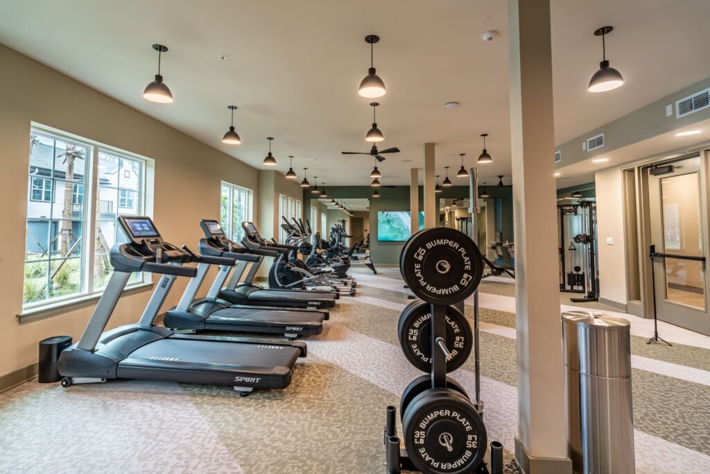 A modern gym with treadmills, rowing machines, and weight racks. Large windows line one wall, and overhead lights illuminate the space.