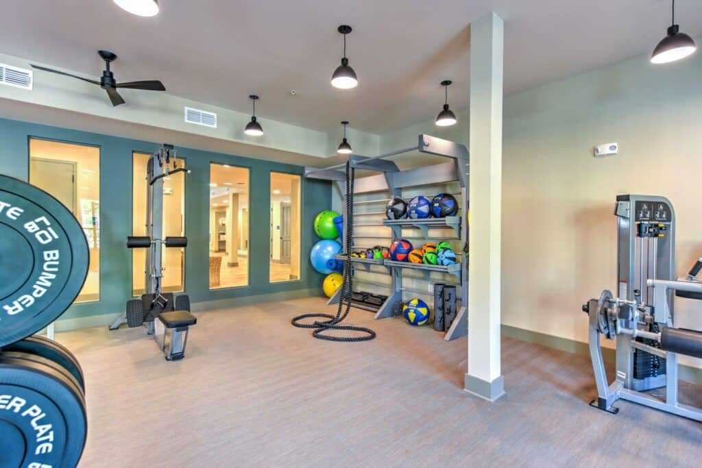 A gym room with exercise equipment, including weights, ropes, a weight machine, and shelves holding balls and kettlebells.