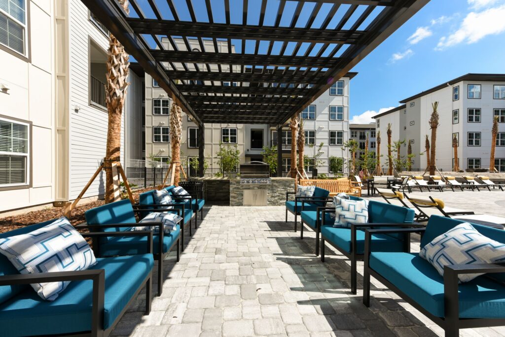 Modern outdoor patio with blue cushioned seating, a pergola, and a grill area. Surrounded by apartment buildings and palm trees under a clear sky.