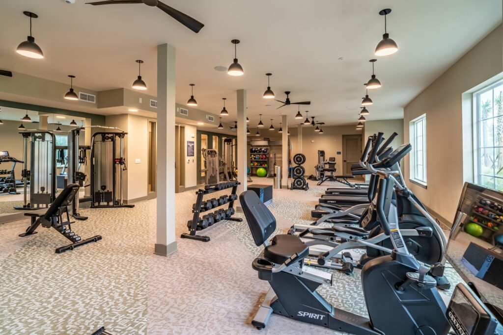 A gym with exercise bikes, weight machines, dumbbells, and fitness balls. The room has large mirrors, ceiling fans, and windows for natural light.