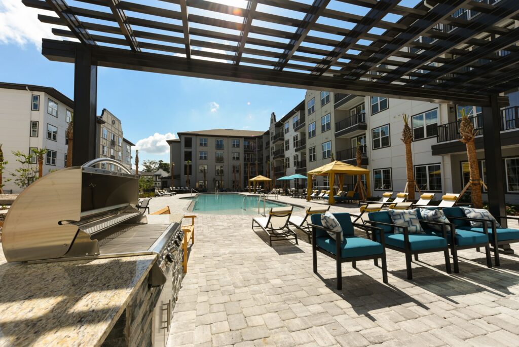 Outdoor pool area with sun loungers, a barbecue grill, and pergola. Surrounded by apartment buildings under a clear sky.