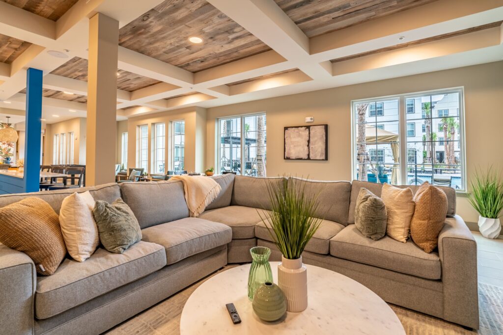 A modern living room features a large sectional sofa with assorted pillows, a round coffee table with vases, and multiple windows allowing natural light to fill the space.