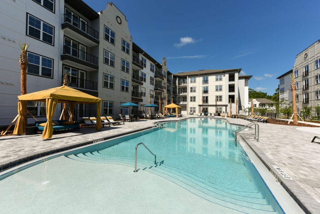 Outdoor pool area surrounded by multi-story apartment buildings, with poolside lounge chairs and cabanas.