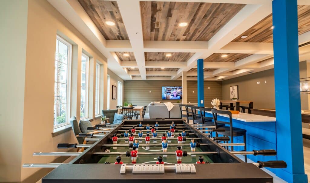 A modern recreational room with a foosball table in the foreground, a sitting area with a TV, and bar seating along a blue counter.