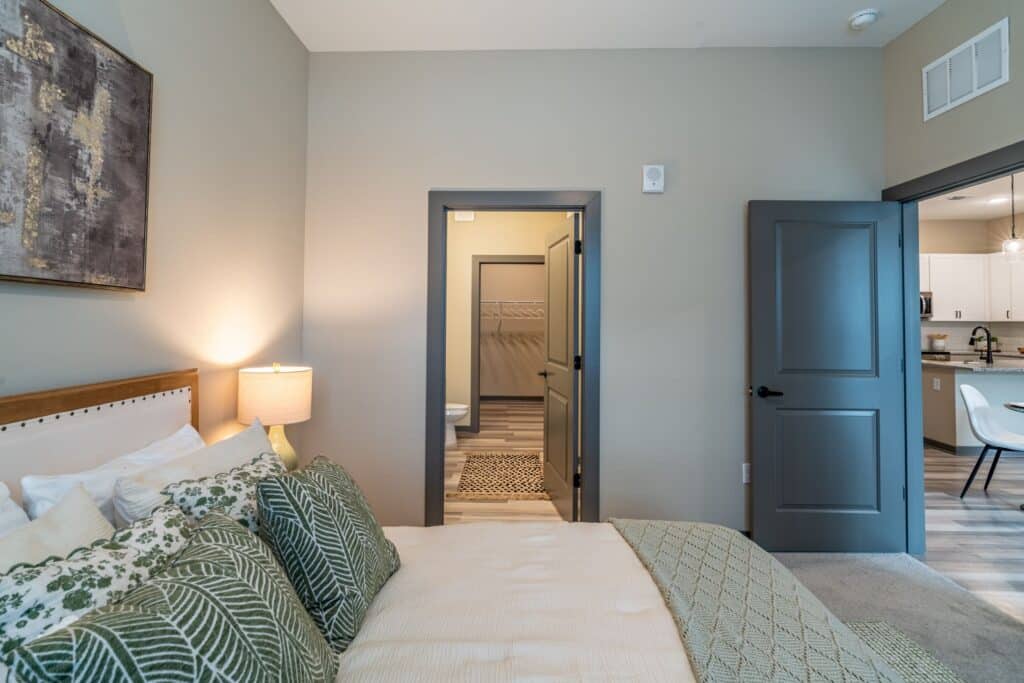 A cozy bedroom with a neatly made bed, green patterned pillows, and a lamp. There is a door open to a walk-in closet, and another door to a kitchen area with white cabinets.