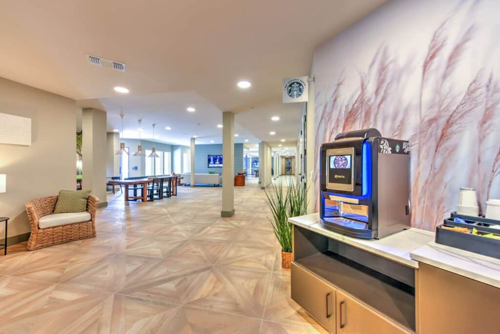 Modern lounge area with a coffee machine on a countertop, wicker chair, long table, and TV screens in the background. Decor includes plant and wall art.