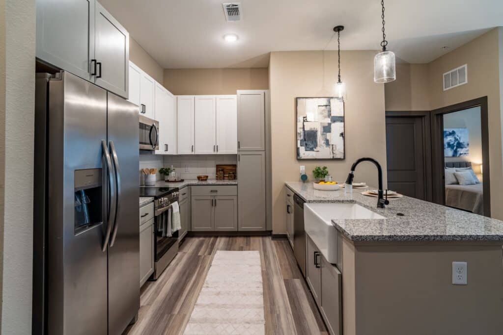 Modern kitchen with stainless steel appliances, white and gray cabinets, and a granite island countertop. Pendant light hangs above the island. A bedroom is visible through an open door.