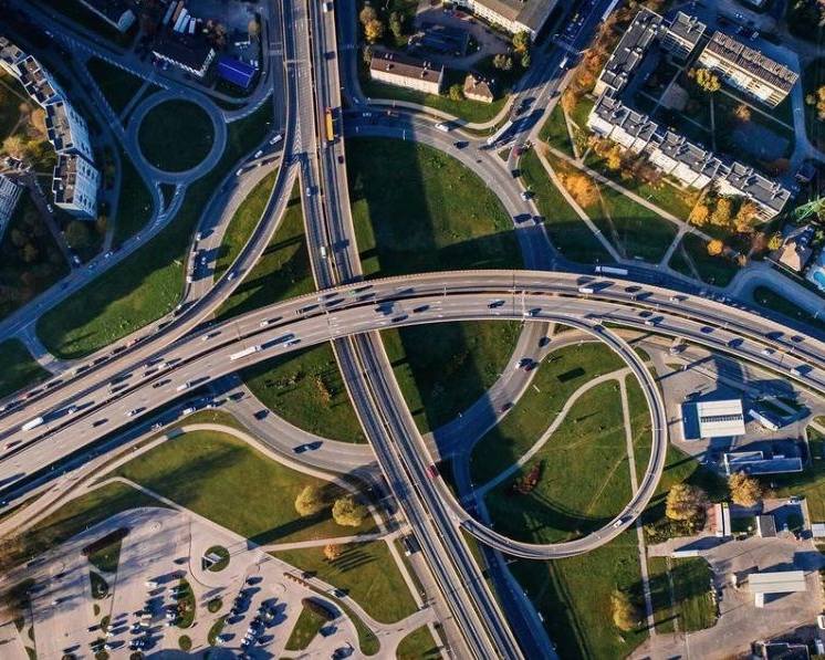 Aerial view of a highway interchange with multiple overpasses and loops, surrounded by greenery and buildings. Vehicles are visible on the roads.