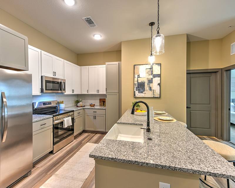 Modern kitchen with stainless steel appliances, white cabinets, and a granite island with two pendant lights. Artwork hangs on the wall above a countertop with fruit and plates.