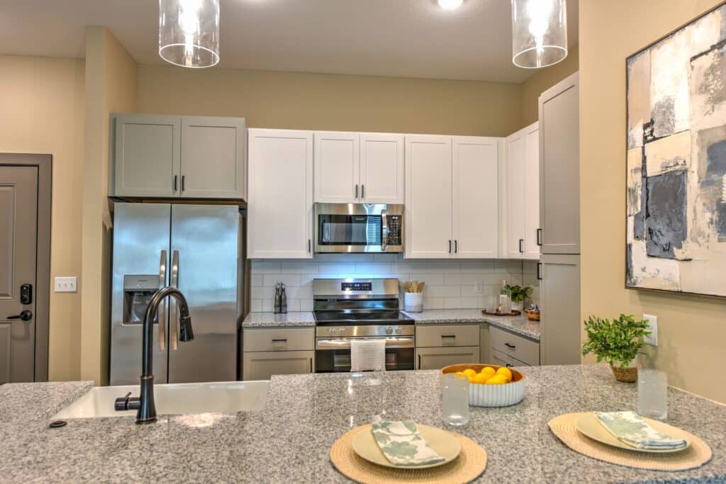 Modern kitchen with stainless steel appliances, white cabinets, granite countertops, and a center island set for two. Artwork hangs on the wall, and a bowl of oranges is on the counter.