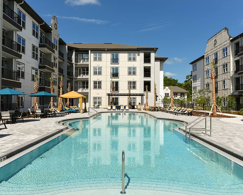 Outdoor swimming pool surrounded by multi-story apartment buildings, with lounge chairs and umbrellas on the deck.