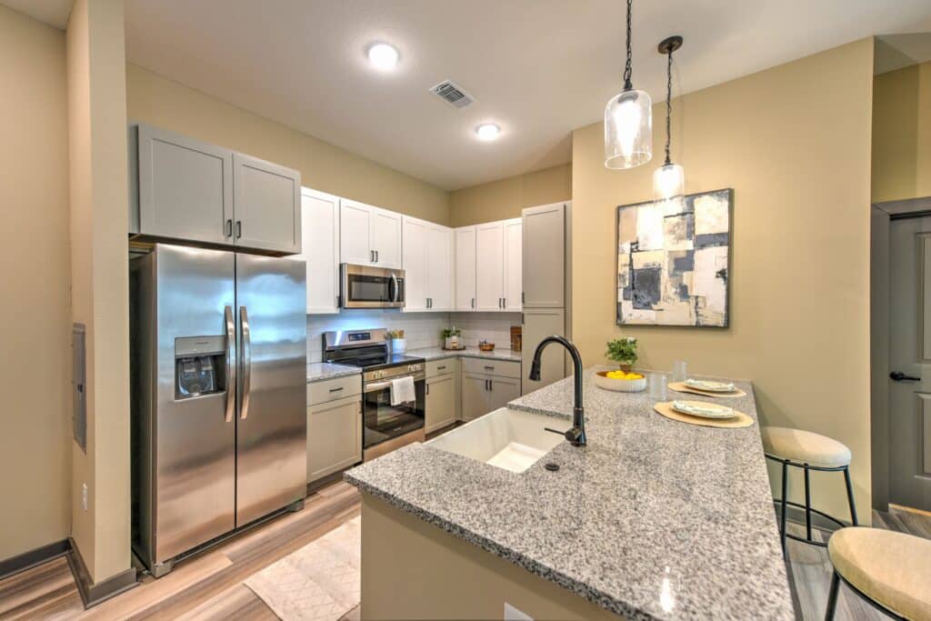 Modern kitchen with granite countertops, stainless steel appliances, pendant lights, and bar seating. Light brown and white cabinets contrast with the beige walls. Abstract art hangs on the wall.