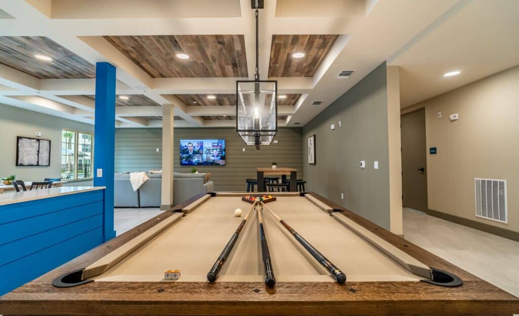 A pool table centered in a modern room with a TV, sofas, and tables in the background. The ceiling features wooden accents and a geometric light fixture hangs above the table.