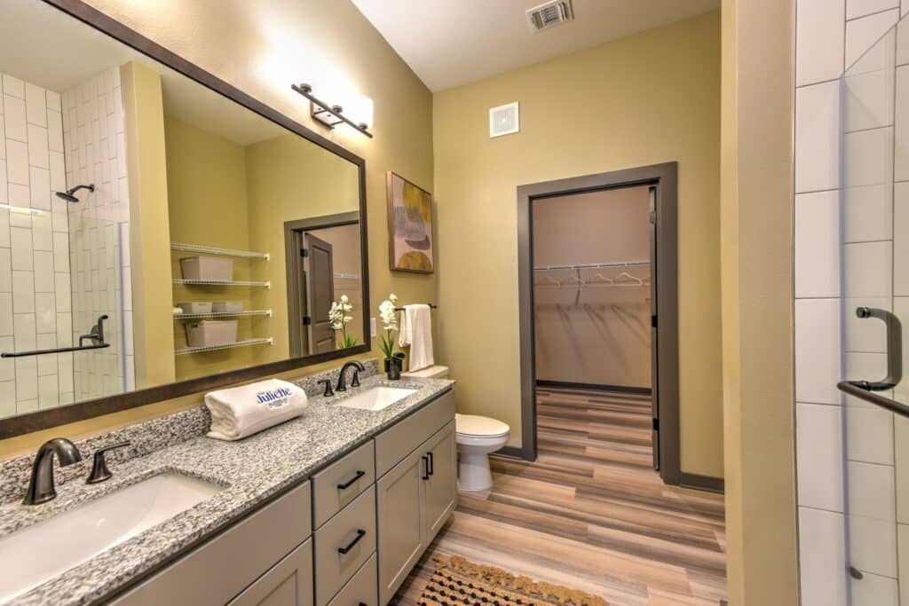 Modern bathroom with dual sinks, granite countertop, large mirror, and beige walls. Includes a toilet, open closet, shower with glass door, and wood-patterned flooring.