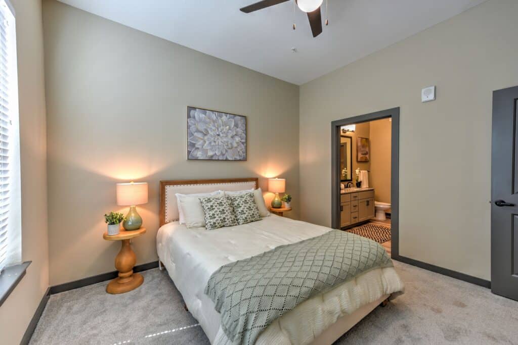 A cozy bedroom with a double bed, two nightstands, wall art, and a view of a connected bathroom. Soft lighting and neutral tones create a calm atmosphere.