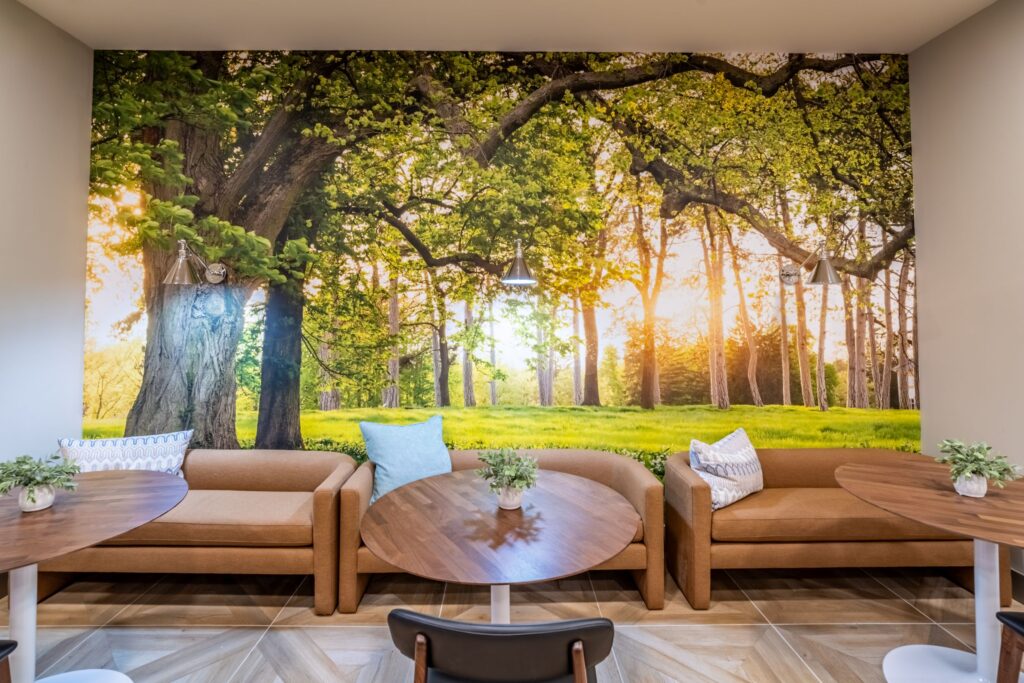 Modern lounge with brown sofas and round wooden tables, featuring a large wall mural of a sunlit forest scene with vibrant green trees.