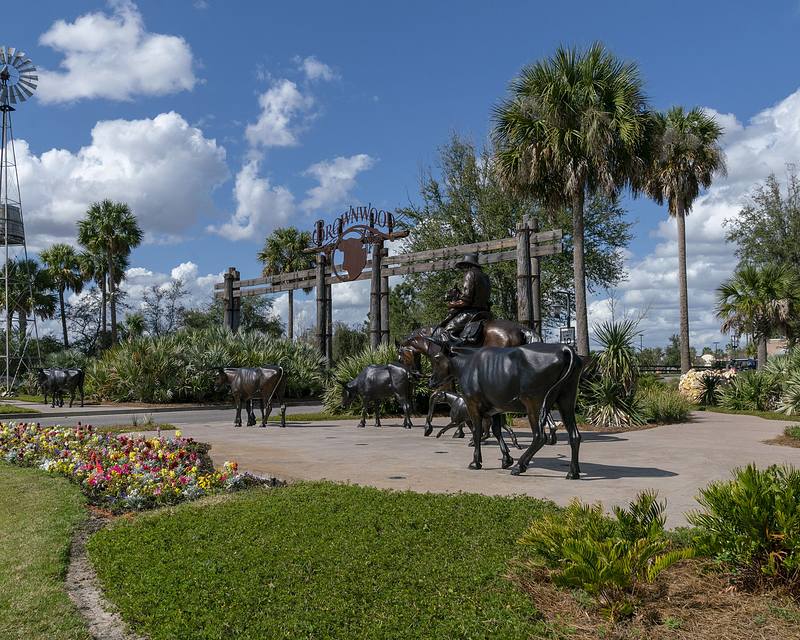 Bronze sculptures of cattle and a cowboy among palm trees and flowers, with a wooden archway and windmill in the background.