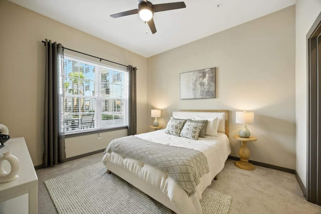 A neatly made bed with pillows and throw blanket sits between two lamps in a beige bedroom with a large window and neutral decor.