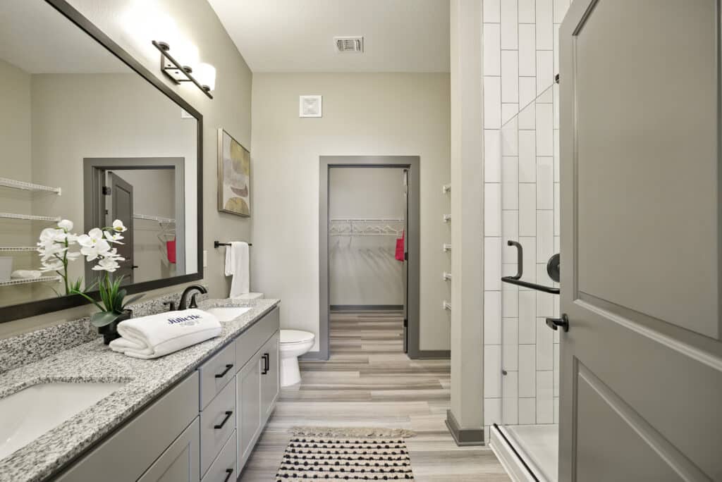 Modern bathroom with gray cabinets, granite countertop, large mirror, walk-in shower, and a view into a walk-in closet. A white orchid and towels are on the counter.