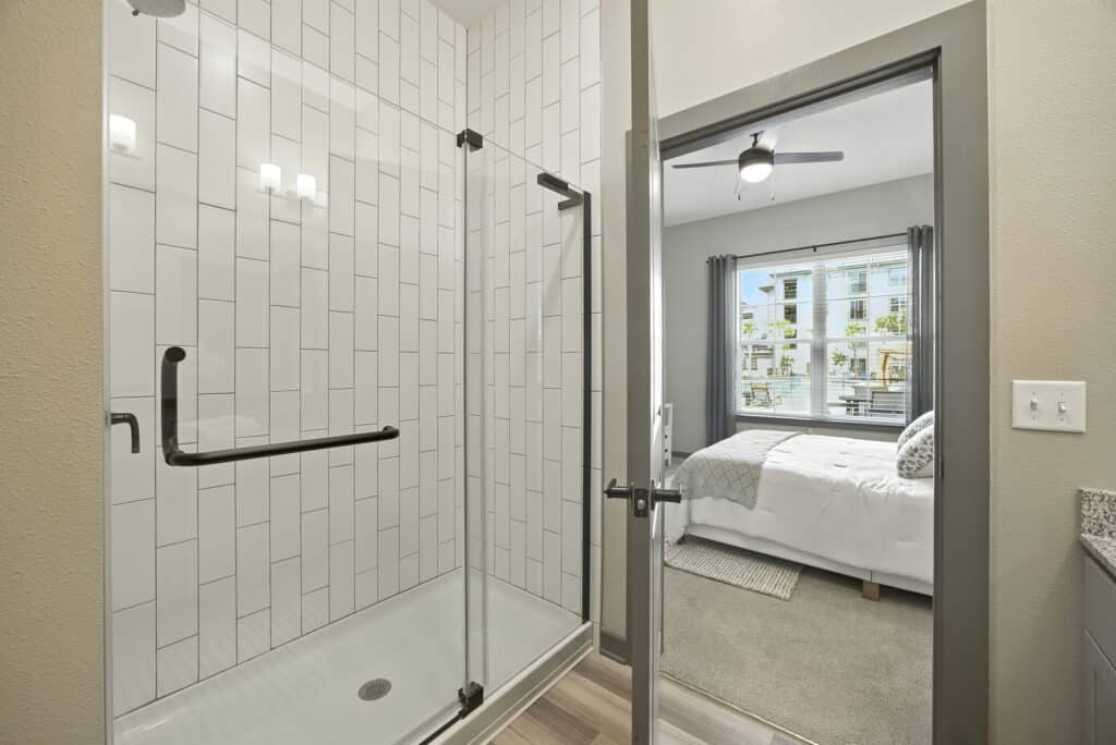 A modern bathroom with a glass shower enclosure and white tile walls, next to a bedroom with a bed, window, and ceiling fan.