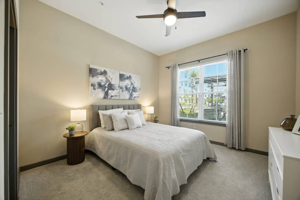 A modern bedroom with a neatly made bed, two bedside tables with lamps, abstract art above the headboard, a window with curtains, and a ceiling fan.