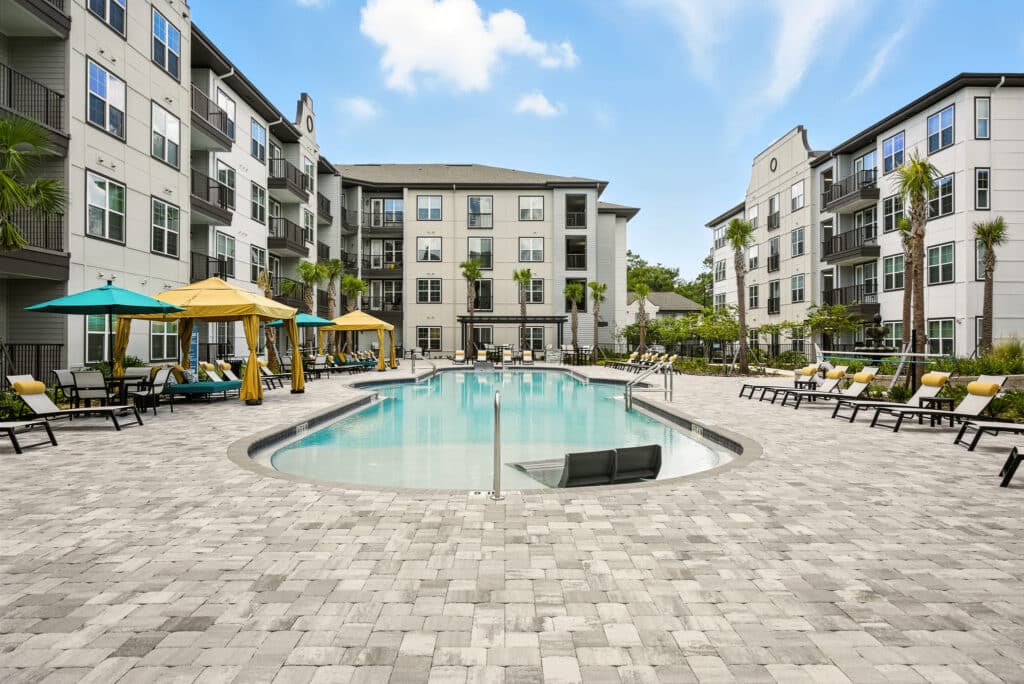 Modern apartment complex courtyard with a central outdoor swimming pool, lounge chairs, and cabanas, surrounded by multi-story residential buildings under a blue sky.