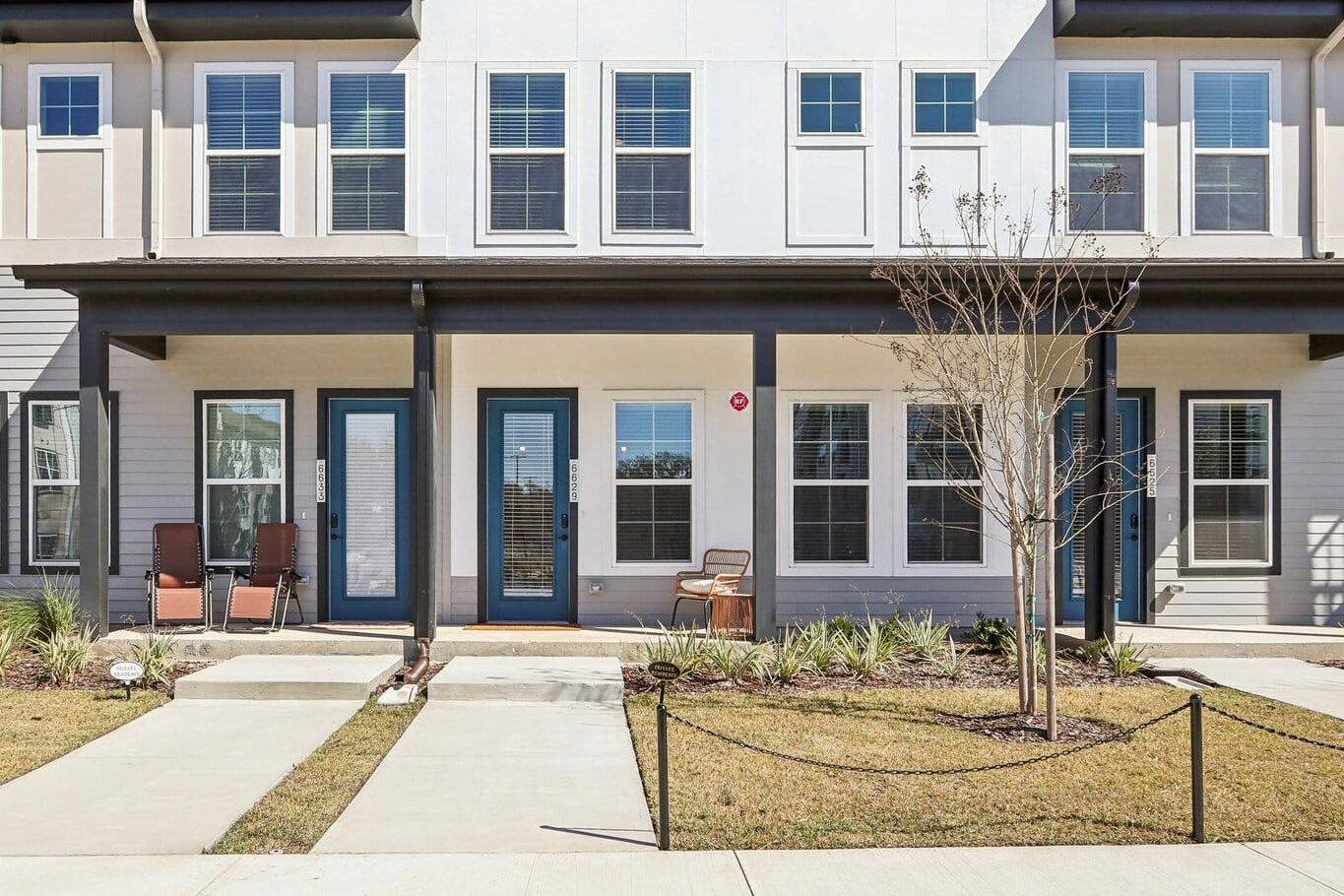 Modern two-story Signature Series Townhomes - The Juliette feature large windows, three front doors, small front patios with chairs, a landscaped yard, and a clear blue sky.