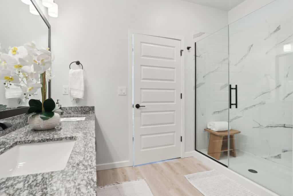 Modern bathroom in Signature Series Townhomes - The Juliette features a granite countertop, double sinks, white door, glass shower enclosure, wooden shower stool, and elegant white orchid decor on the counter.